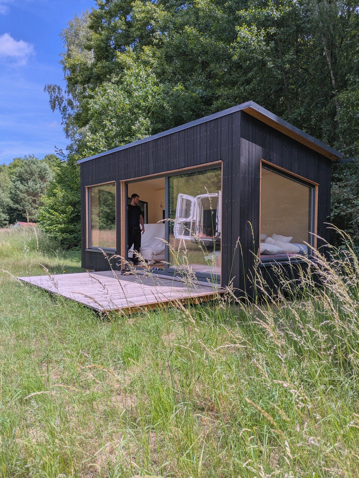 Schwarz geschalter LandAnker mit Glasfront und Holzdeck in hohem Gras, drinnen ein gemachtes Bett