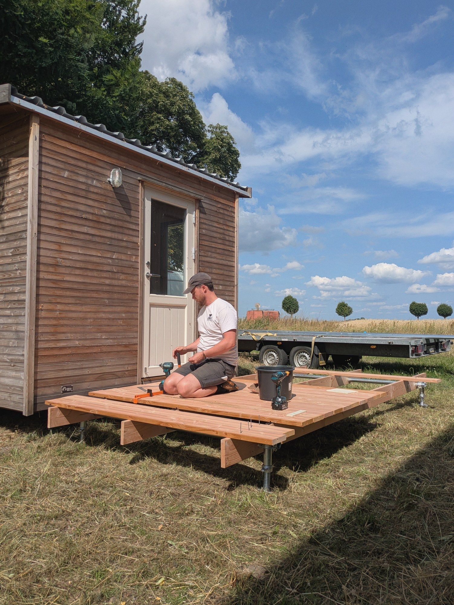 Handwerker schraubt auf einem neuen Holzpodest vor einem verwitterten LandAnker, dahinter der leere Anhänger